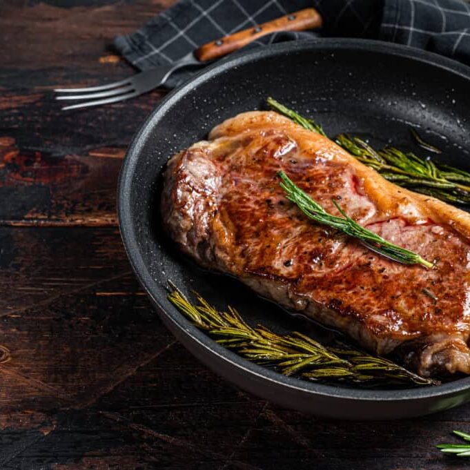 Grilled new york strip beef meat steak in a pan with herbs. Dark wooden background. Top view. Copy space.