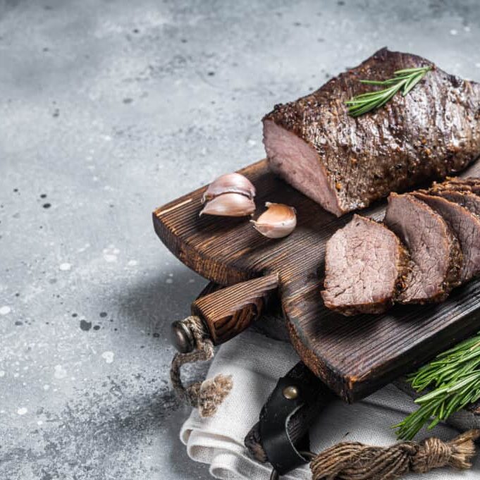 Roast and sliced tri tip beef steak on a wooden board with herbs. Gray background. Top view. Copy space.